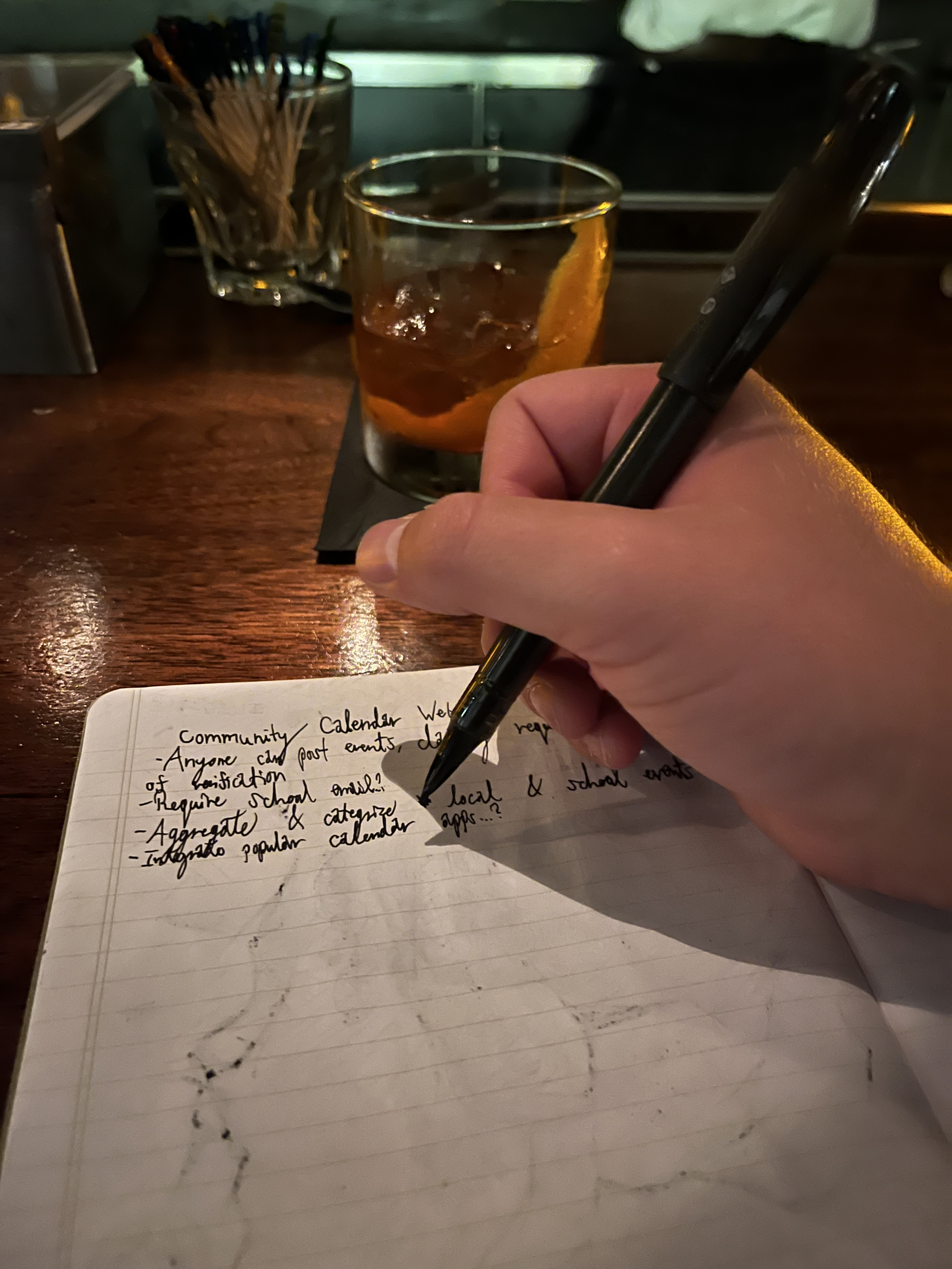 A hand writing in a notebook with a fountain pen. A cocktail is in the background on a wooden table